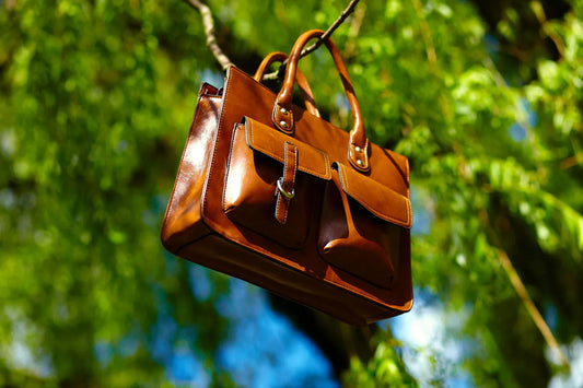 Brown leather satchel handbag with front pockets hanging from a tree branch in a forest setting.