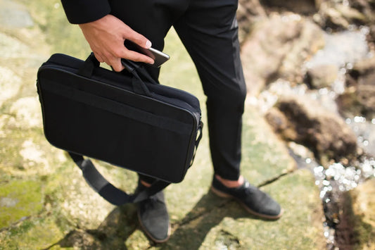 A man in black pants and shoes holds a black fabric briefcase and a smartphone while standing on mossy rocks by the ocean.